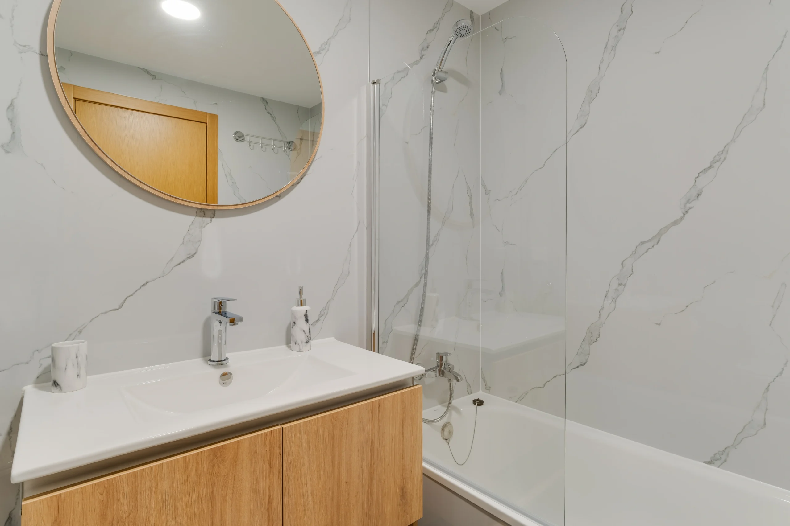 Łazienka z wanną, przeszklonym parawanem i okrągłym lustrem, jasne kafle.